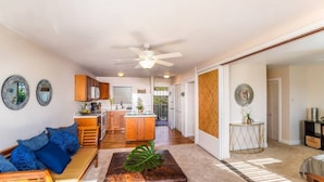 A spacious living area with a sofa, ottoman, and a dining table. There is a kitchen in the background with wooden cabinets and a refrigerator.