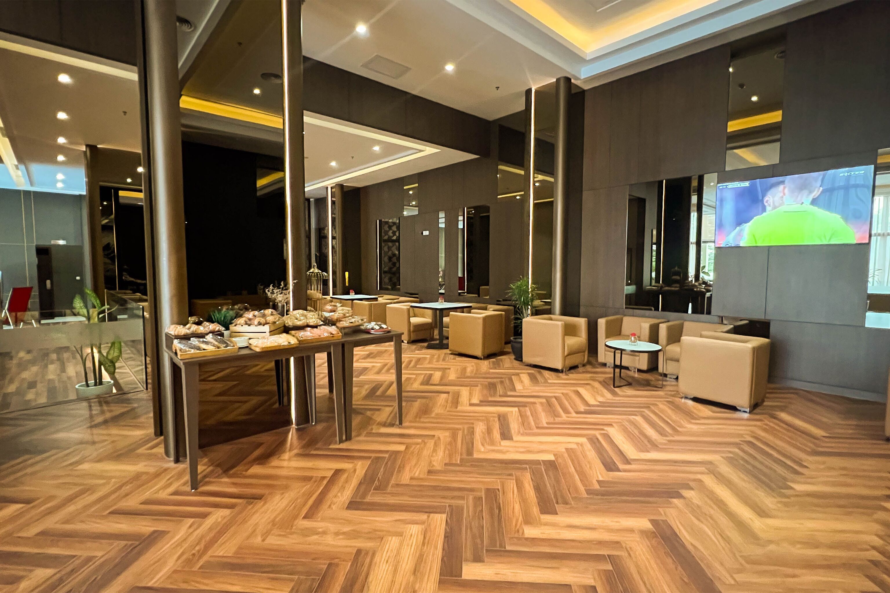 Un lobby espacioso con piso de madera en patrón de espiga, muebles modernos y una pantalla grande mostrando a una persona con camisa verde.