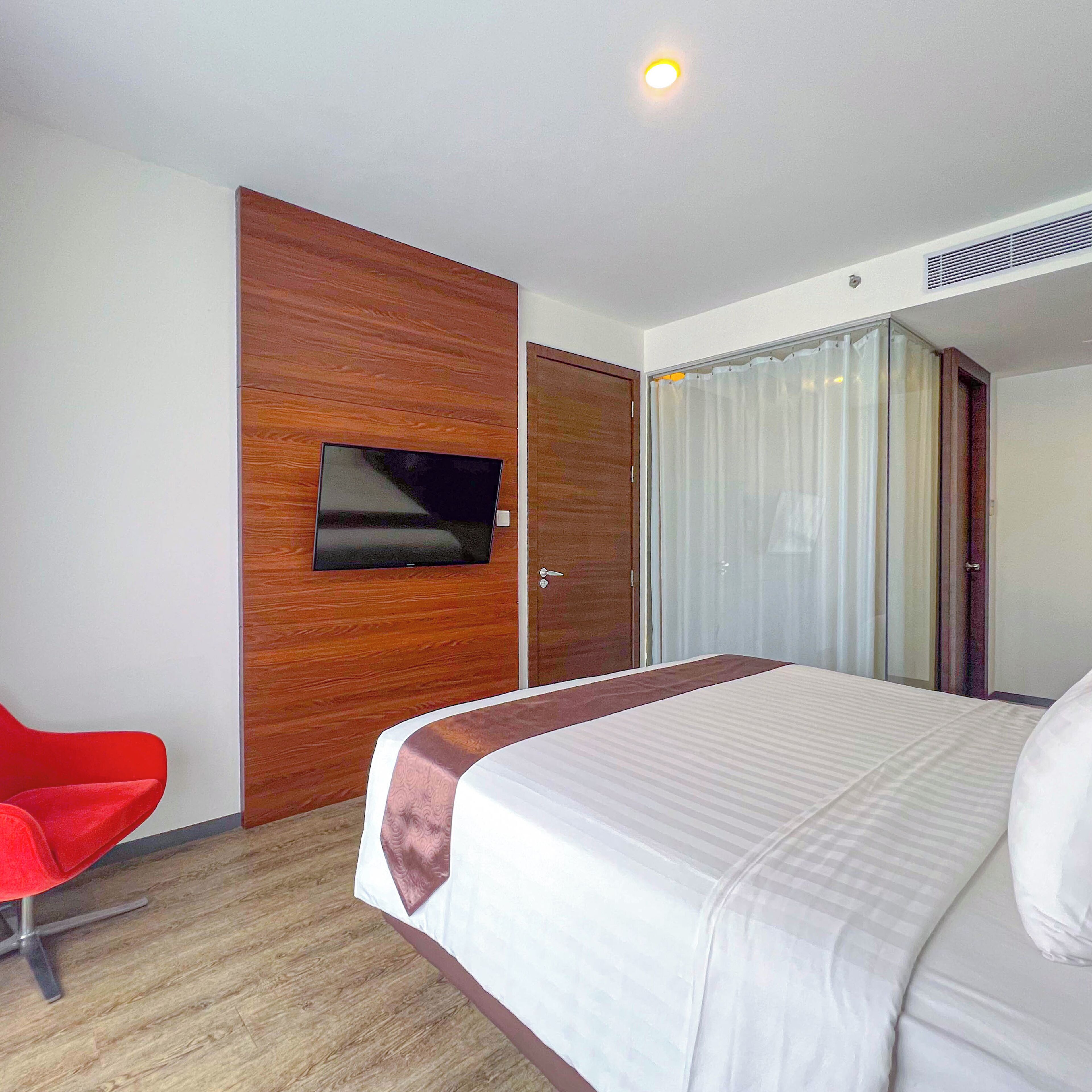 A hotel room with a bed, a TV mounted on a wooden panel, a red chair, and a door with a curtain.