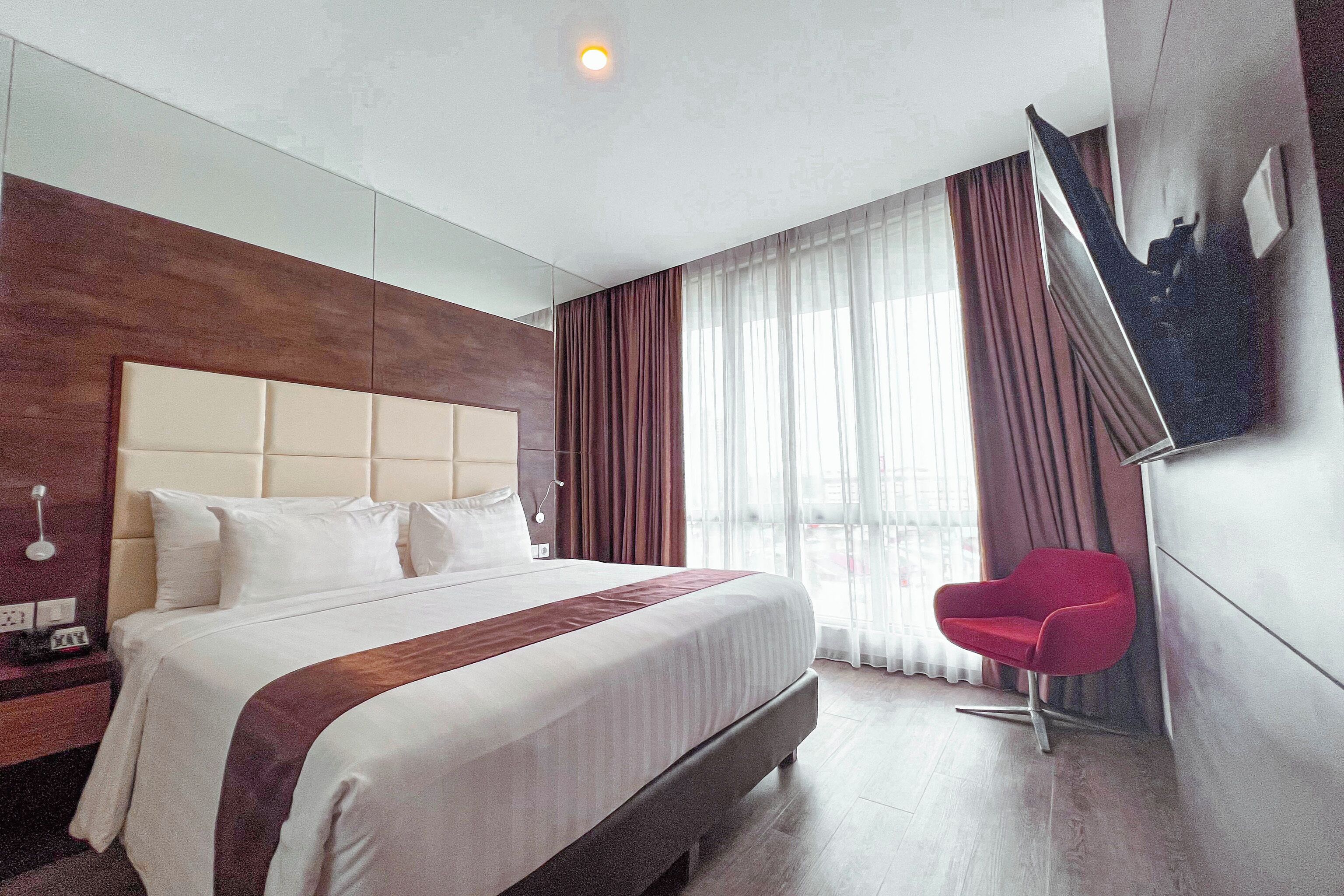 Ett hotellrum med en stor säng, en röd fåtölj och utsikt över staden genom fönstret.