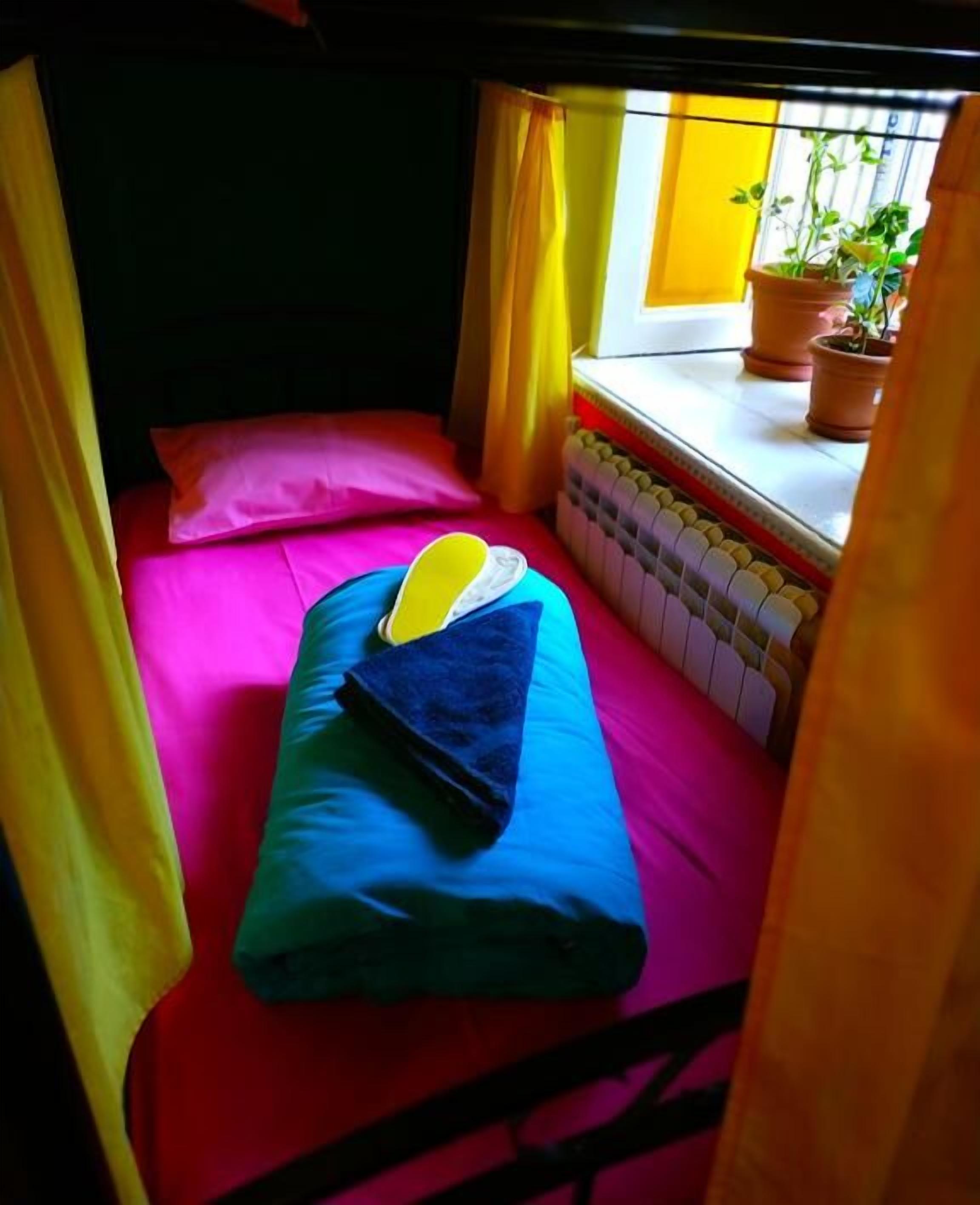 A small, cozy room with a single bed, bright yellow curtains, and a window with potted plants.