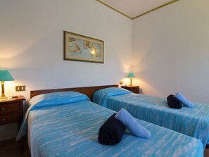 Ein Hotelzimmer mit zwei Betten, jeweils mit blauer Bettdecke und Kissen. Zwei Nachttische mit Lampen sowie ein gerahmtes Bild an der Wand.