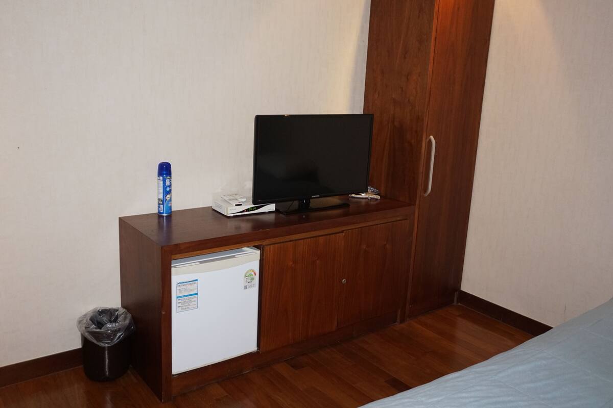 Une petite pièce comprenant un meuble en bois, une télévision, un mini-réfrigérateur et une poubelle.
