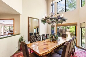 A dining area with a large wooden table, upholstered chairs, a chandelier, and a large mirror.