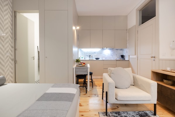 A compact hotel room with a bed, a sofa, a small kitchen area, and a dining table.