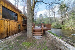 A wooden cabin with a stone wall and a wooden staircase leading to a deck overlooking a river.