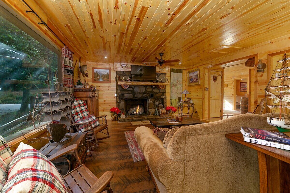 A cozy cabin interior with a stone fireplace, wooden furniture, and a large window overlooking a forested area.