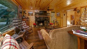 A cozy cabin interior with a stone fireplace, wooden furniture, and a large window overlooking a forested area.