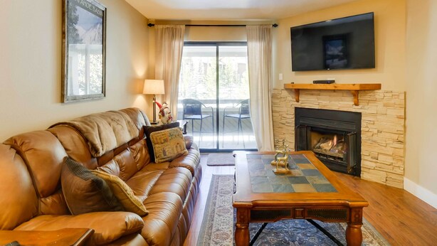 A living room with a leather sofa, a coffee table, a fireplace, a TV mounted on the wall, and a large window with curtains. - Chic & Cozy Breck Condo: Ski-In/Ski-Out at Peak 8! (Breckenridge)