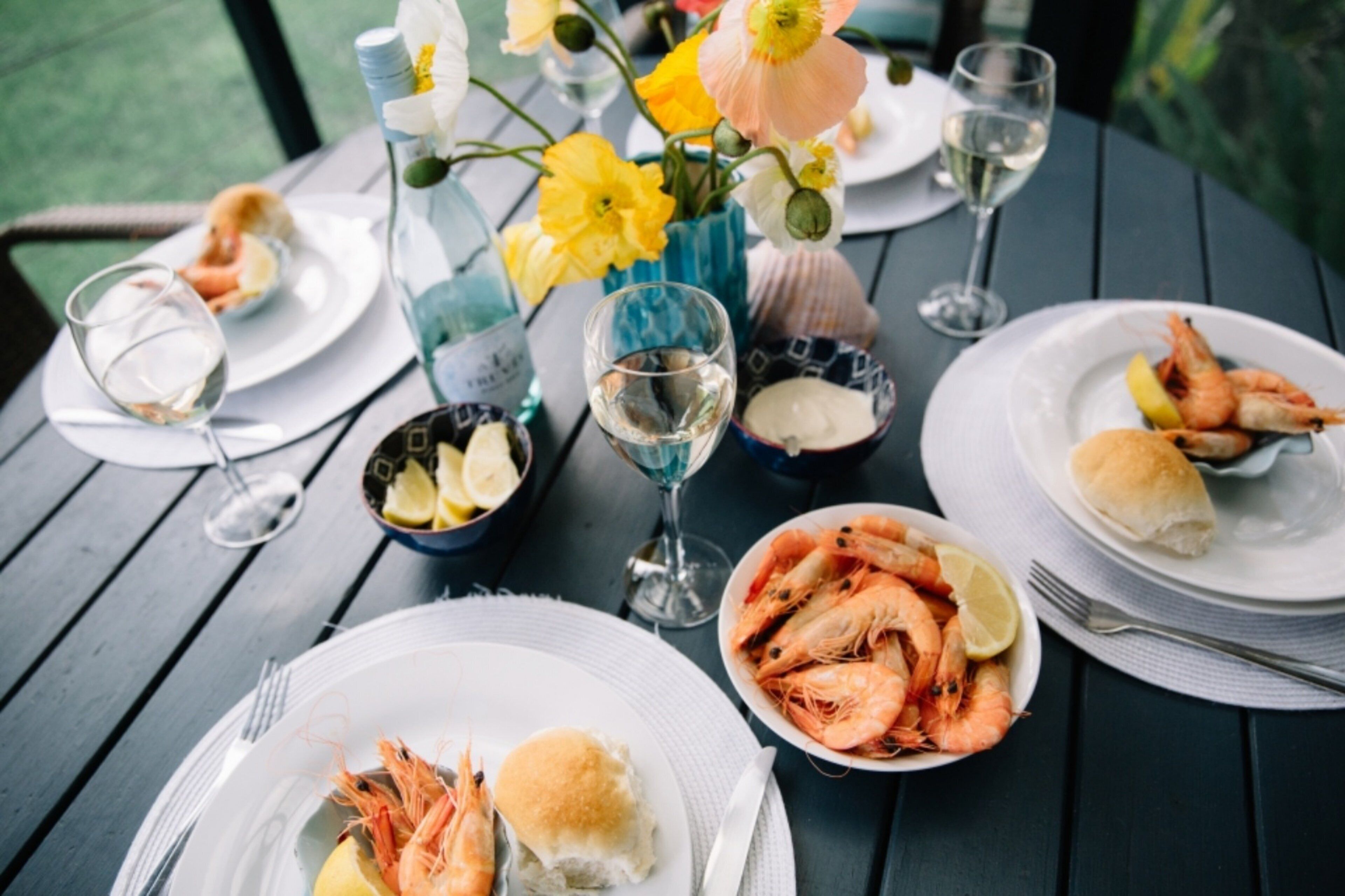 A table set for a meal with plates of shrimp, glasses of white wine, and a vase with yellow flowers.