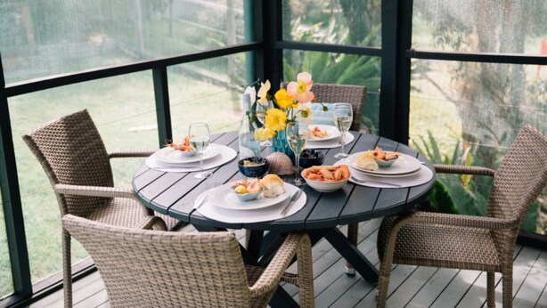A round table with wicker chairs on a wooden deck, surrounded by glass walls with a view of greenery. - Salt Breeze - upstairs only (Lake Cathie)