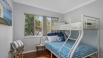 Uma cama beliche com edredom estampado em azul e branco, uma mesinha de cabeceira, uma cadeira e uma janela com vista para árvores.