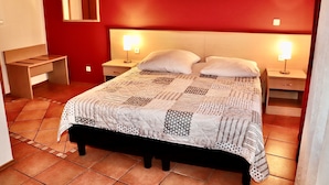 A neatly made bed with a patterned bedspread, two bedside lamps, a nightstand, a wooden bench, and a red wall with a mirror and artwork. - Hotel Romina (Frankfurt)