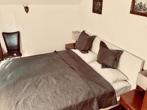 A neatly made bed with a patterned bedspread, a bedside table with a lamp, and a chair in a room with plain walls and minimal decor. - Hotel Romina (Frankfurt)