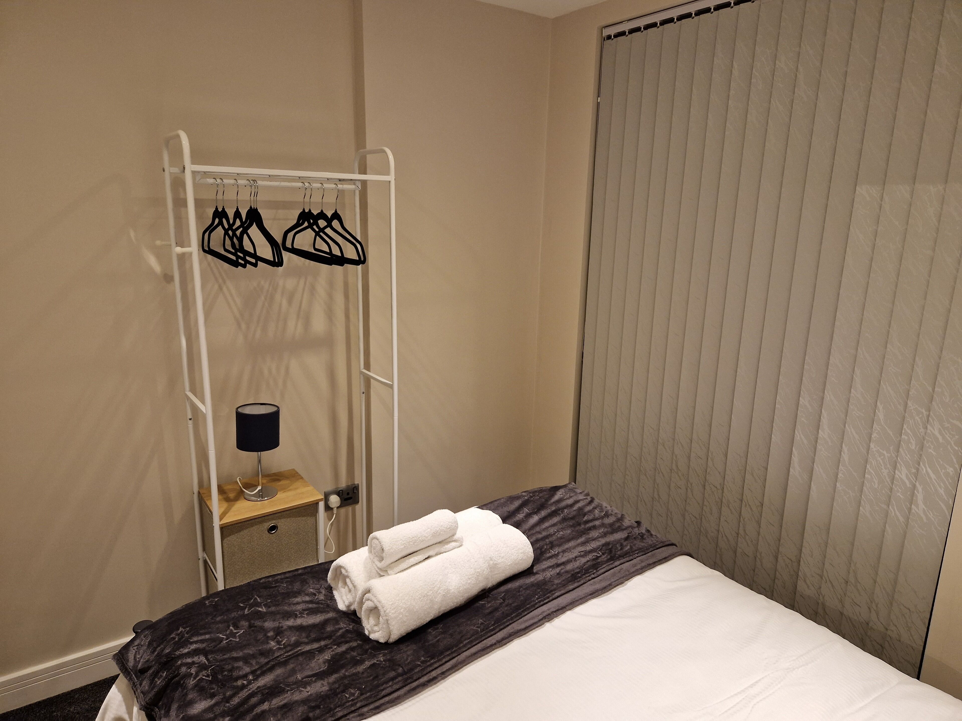 A hotel room with a bed, a towel rack with hangers, a small table with a lamp, and a window with blinds.