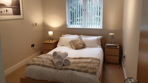 Une chambre à coucher comprenant un lit, deux tables de chevet, une fenêtre avec des stores, un tableau accroché au mur et un radiateur.