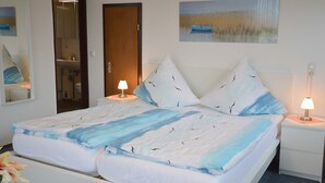 Ein ordentlich bezogenes Doppelbett mit blauer und weißer Bettdecke, zwei Nachttische mit Lampen, eine Holztür, ein durch eine geöffnete Tür einsehbarer Badezimmerbereich und ein Bild eines Bootes auf dem Wasser an der Wand.