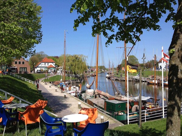 Un café situé au bord de la rivière, avec une terrasse, un quai où sont amarrés des bateaux et un bâtiment surmonté d’un toit rouge.