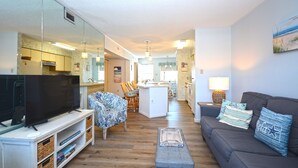 A living room with a television, sofa, and ottoman. There is a kitchen area in the background with a dining table and chairs. - Nice 2 bedroom condo with pristine hardwood floors, free WiFi, and an outdoor pool located midtown and only a block to the beach! (Ocean City)