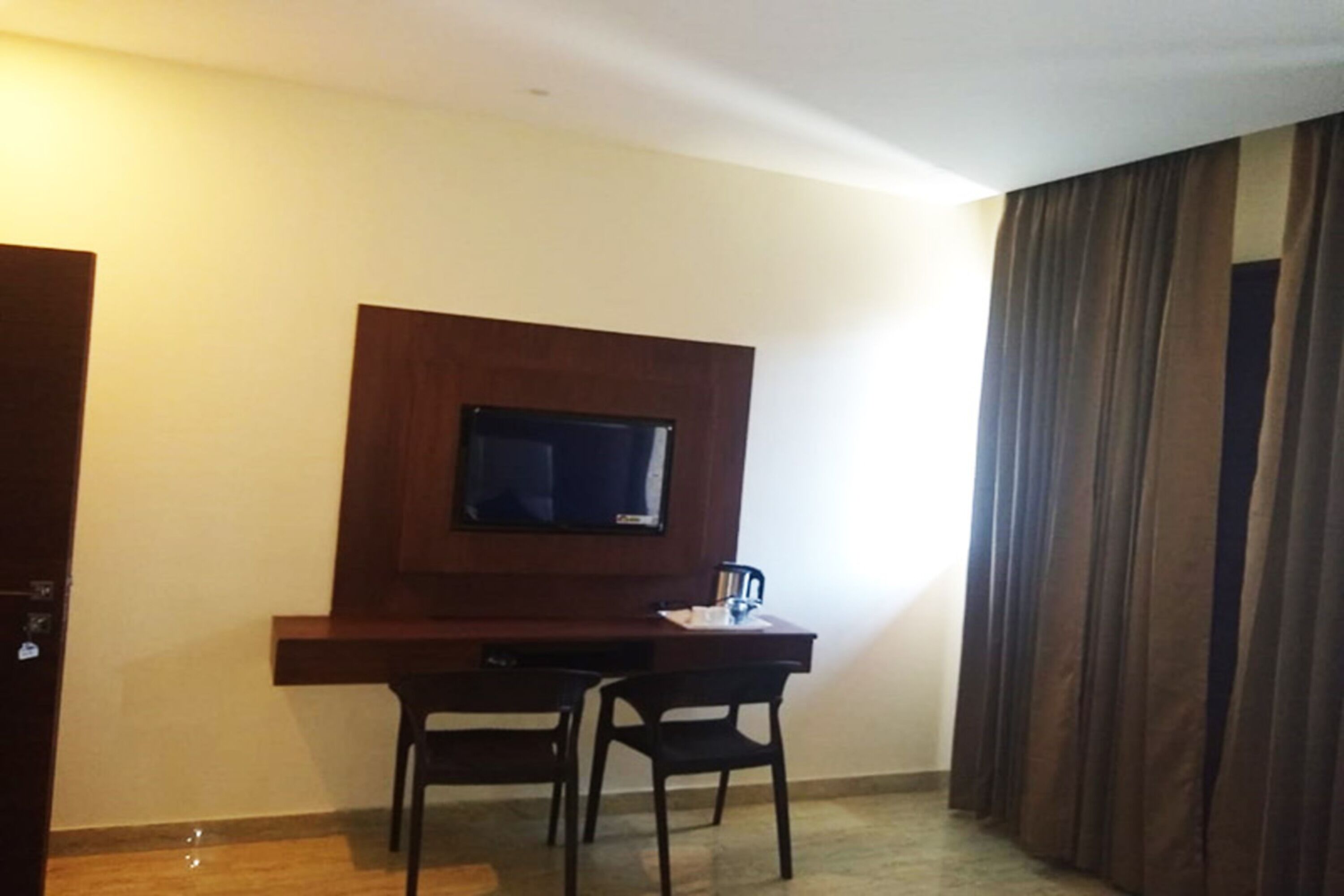 A room with a wooden desk, two chairs, a flat-screen TV mounted on the wall, and a large window with curtains.