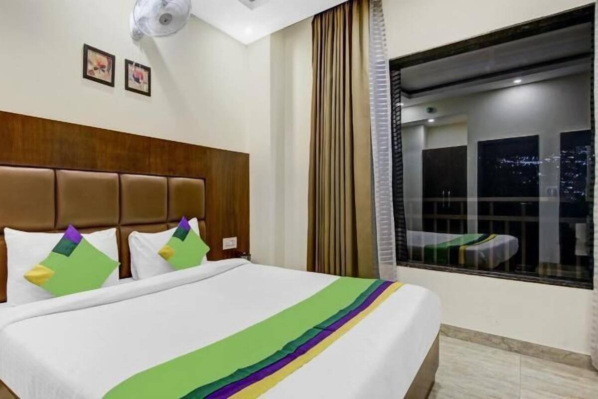 A hotel room with a bed, a headboard, a window with curtains, and a wall-mounted air conditioner.