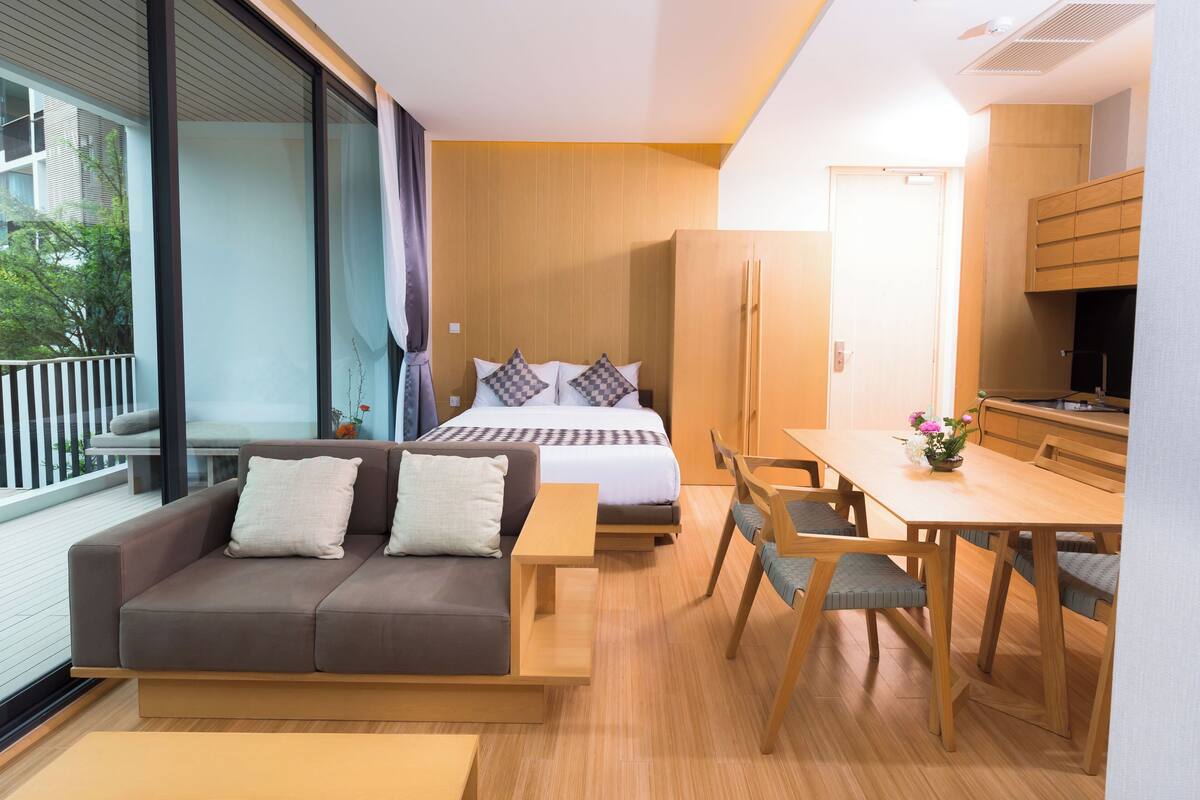 A modern hotel room with a bed, a sofa, a dining table, and a kitchen area.