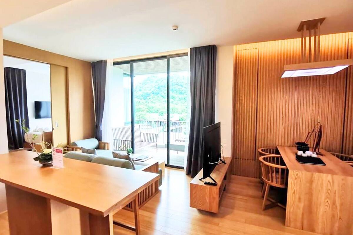A modern hotel room with a wooden dining table, a sofa, a TV, and a large window offering a view of the outdoors.