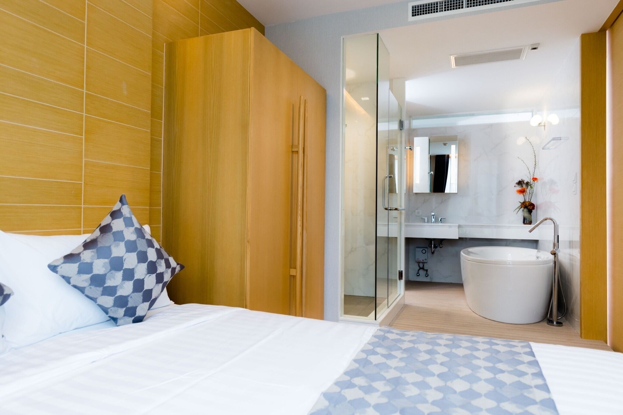 A modern hotel room with a bed, a checkered pillow, a glass shower enclosure, a white sink, and a large bathtub.