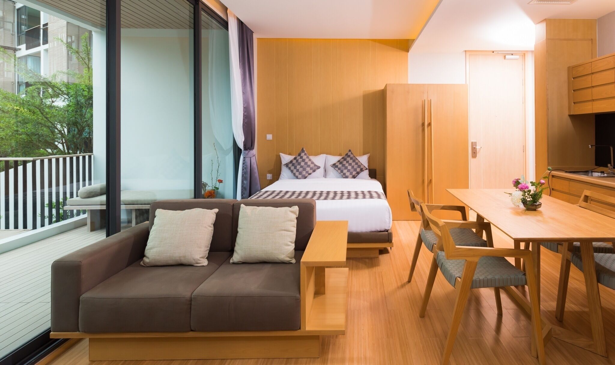 A modern hotel room with a balcony, a sofa, a bed with checkered pillows, a wooden dining table, and a kitchen area.
