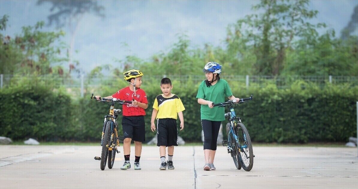 Three people with bicycles on a paved surface, surrounded by greenery.