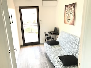 A room with a patterned sofa, a desk with a laptop, a chair, and a door leading to an outdoor area.