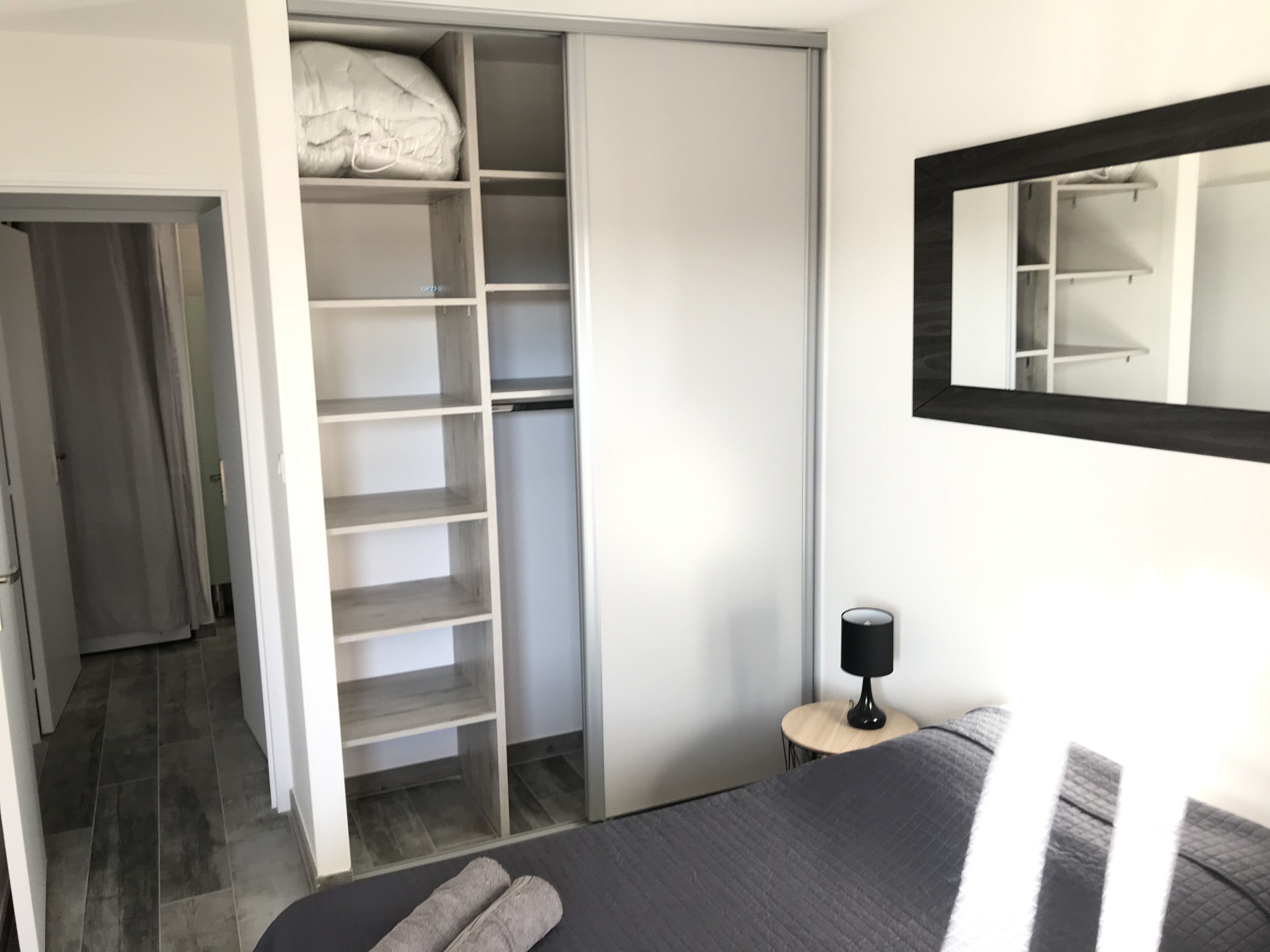 Ein Schlafzimmer mit Bett, Kleiderschrank, einem kleinen Tisch mit Lampe und einem Spiegel an der Wand.