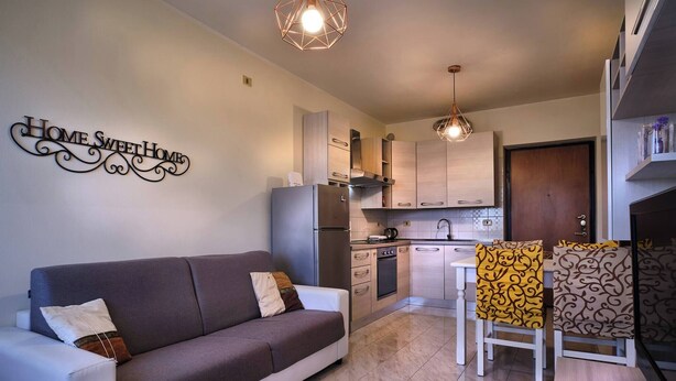 A modern living room with a sofa, a dining table with chairs, a kitchen area with cabinets and appliances, and decorative wall art.