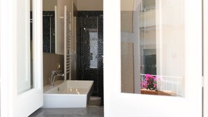 A modern bathroom with a bathtub, a shower, and a toilet. There is a balcony with a railing and a potted plant visible through the window. - Palazzo Gargano (Amalfi)