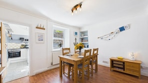 A compact kitchen with a built-in oven, a dining area with a wooden table and chairs, and a small sideboard. - Shell Cottage 2 Bedroom Holiday (Tenby)