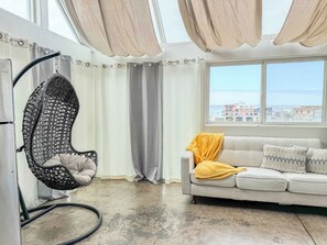 A modern living room with a hanging chair, a sofa with a yellow throw, and a view of buildings through a large window. - 303/C Little Italy double balcony Loft /Parking (San Diego)