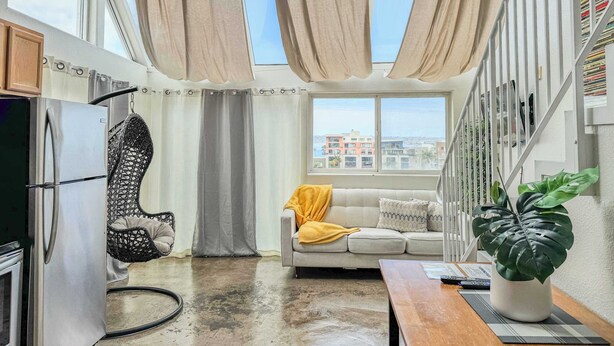 A modern living room with a sofa, a coffee table, a plant, and a view of the city through large windows. - 303/C Little Italy double balcony Loft /Parking (San Diego)