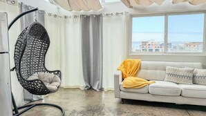 A modern living room with a hanging chair, a sofa with a yellow throw, and a view of buildings through a large window. - 303/C Little Italy double balcony Loft /Parking (San Diego)