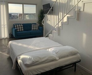 A bedroom with a bed, a blue sofa with checkered pillows, a staircase, and a view of buildings through a window. - 303/C Little Italy double balcony Loft /Parking (San Diego)