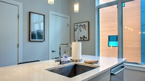 A modern kitchen with a sink, faucet, and countertop. There is a window with blinds and a door in the background.