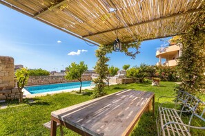 Una mesa de madera y un banco de metal bajo una pérgola de bambú, con una piscina y un muro de piedra al fondo.