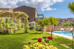 Un patio con piscina, un pequeño jardín con flores y un edificio de techo a la tosca.