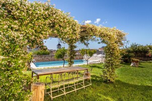 Un área de piscina con una mesa y sillas de madera, rodeada de exuberante follaje y plantas en flor.