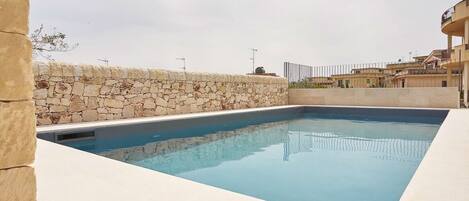A rectangular swimming pool with a stone wall on one side and a white tiled deck on the other.