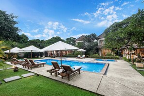 A swimming pool with lounge chairs and umbrellas, surrounded by greenery and buildings. - Villa Pantai Karang (Denpasar)