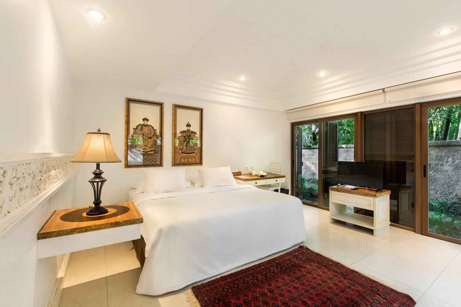 A modern bedroom with a large bed, a bedside table with a lamp, a TV, and a sliding glass door leading to a balcony.