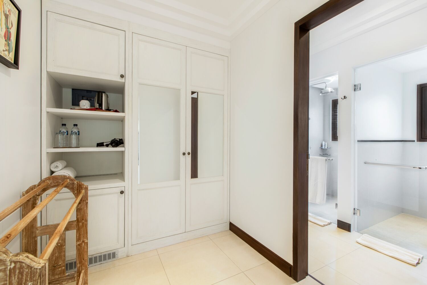 Una habitación con un armario blanco, una silla de madera y un baño visible a través de una puerta de cristal.