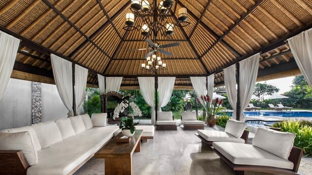 A spacious lounge area with a high thatched ceiling, white cushioned seating, and a view of a pool. - Villa Pantai Karang (Denpasar)