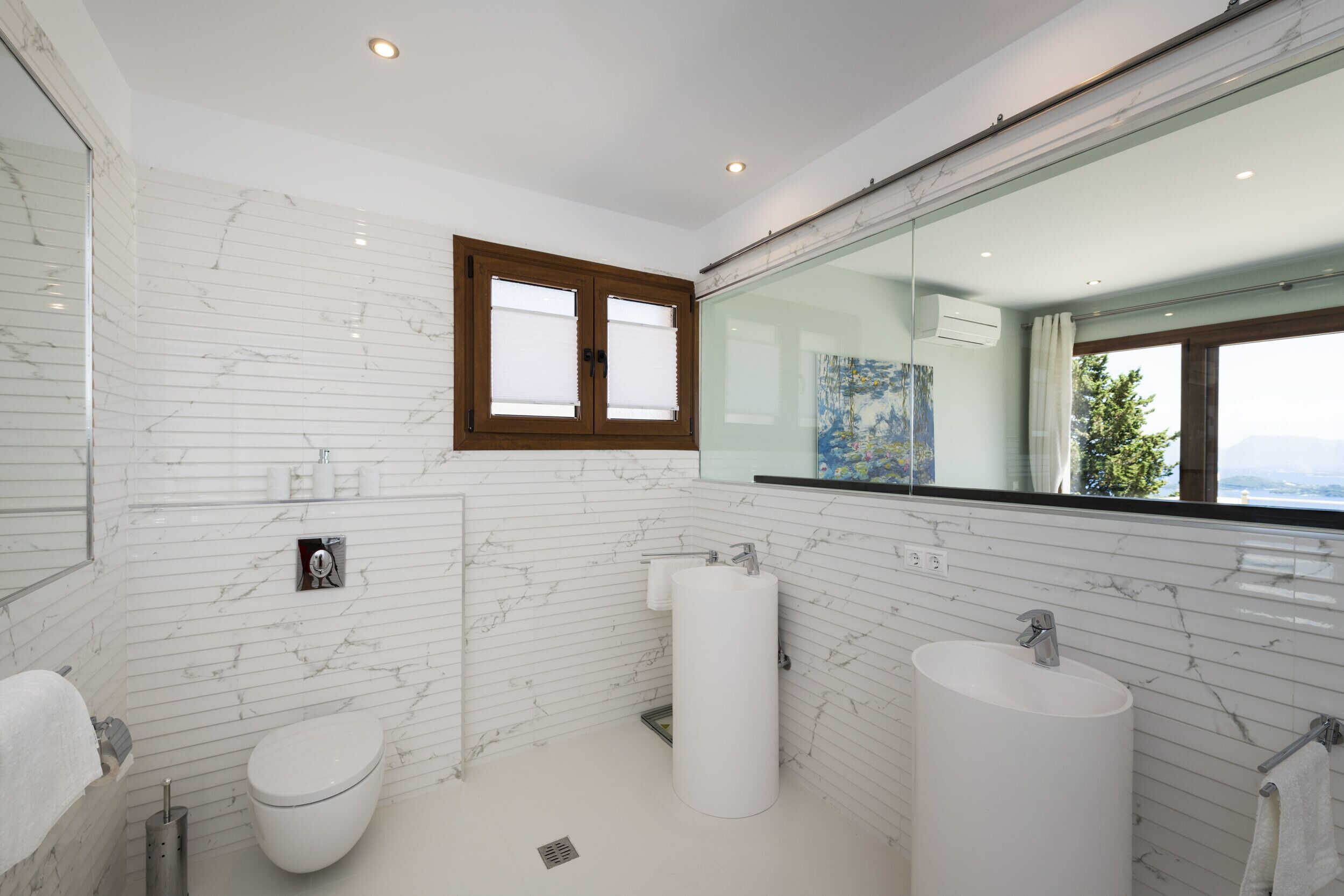Een moderne badkamer met witte marmeren wanden, een grote spiegel, een toilet, een wastafel en een raam met uitzicht.