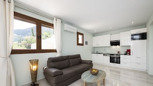 A modern living room with a brown leather sofa, a round coffee table with a bowl, and a floor lamp. A kitchen with white cabinets and appliances is visible in the background. - Secluded THE VIEW Villa ideal for 10 adults & 7 kids w/t pool (Nidri)